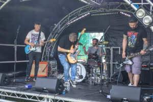 A photo of a band playing on stage at the 2024 Accessible Festival. A guitarist is playing a solo at the centre of the stage, with supporting guitarists at either side. A drummer is keeping rhythm in the background.