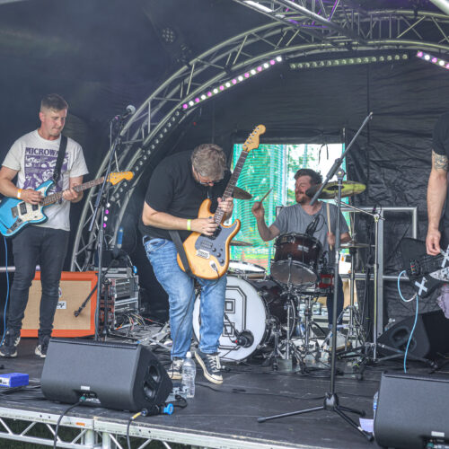 267A7007 A photo of a band playing on stage at the 2024 Accessible Festival. A guitarist is playing a solo at the centre of the stage, with supporting guitarists at either side. A drummer is keeping rhythm in the background.