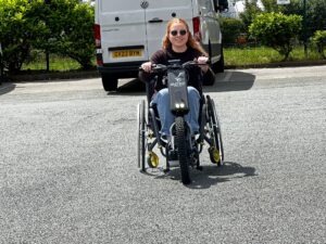 A picture of Kate racing across a car park on a Batec mobility hand bike attached to a wheelchair.