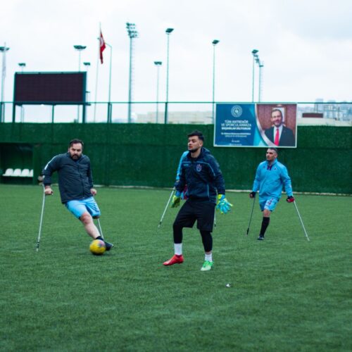 Untitled design-3 Image of accessible sports. Three footballers playing football on a 3g pitch. Two of the footballers have lower leg amputations and are using crutches. The third footballer is wearing goalkeeping gloves and has a prosthetic leg.