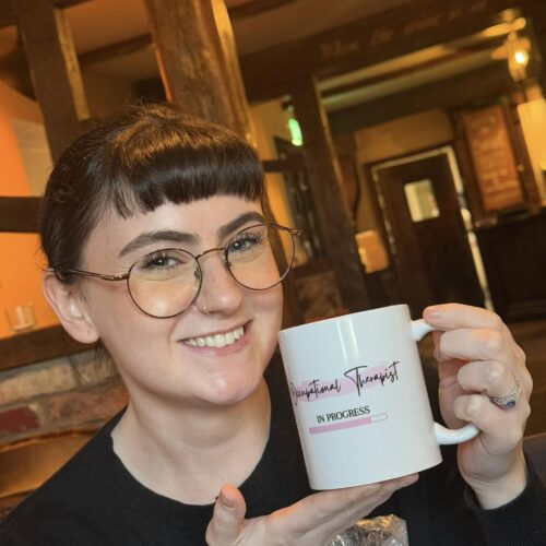 Ciara OT in Progress Picture of Ciara smiling and holding a mug with the caption 'Occupational therapist in progress'.